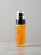 A frosted glass bottle filled with amber-orange liquid, featuring a black pump and a clear cap, standing on a light gray surface against a neutral gray background.